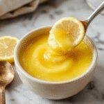 A vibrant jar of bright yellow lemon curd alongside fresh lemons and a spoon, showcasing its creamy texture and zesty color.