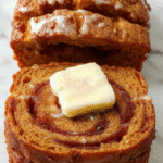 A close-up of a freshly baked pumpkin bread loaf with a cinnamon swirl visible inside, topped with a dusting of powdered sugar and a pat of melting butter. The bread is sliced, revealing its moist texture and beautiful swirl pattern, presented on a rustic wooden board with autumn leaves in the background.