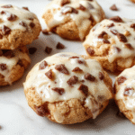 A close-up of freshly baked Samoa cookies arranged on a rustic wooden platter. The cookies are golden-brown with toasted coconut and caramel drizzle on top, with a sticky caramel layer visible beneath the toasted coconut. The background is softly blurred with hints of a baking scene, highlighting the cookies' crispy edges and gooey caramel texture.
